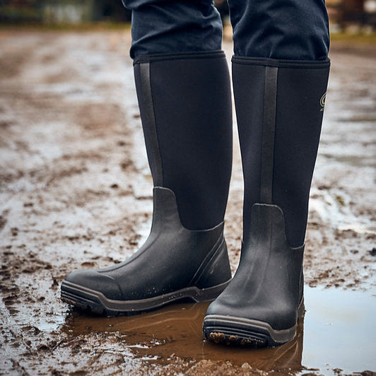Person wearing black rubber boots standing in a muddy puddle.