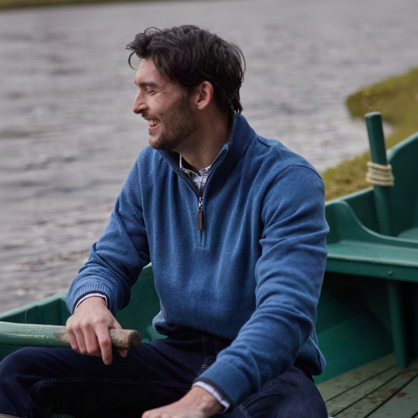 A man wearing a blue quarter-zip sweater and dark jeans sits in a green boat on water, holding an oar.