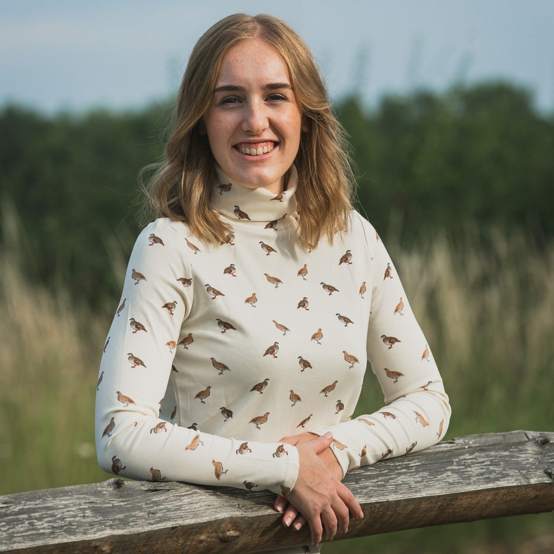 Woman wearing a white turtleneck with bird pattern sitting on a wooden railing outdoors.