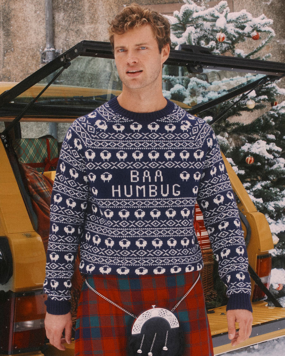 Man wearing a festive sweater with 'BAA HUMBUG' text, standing in front of a vintage car and Christmas tree.