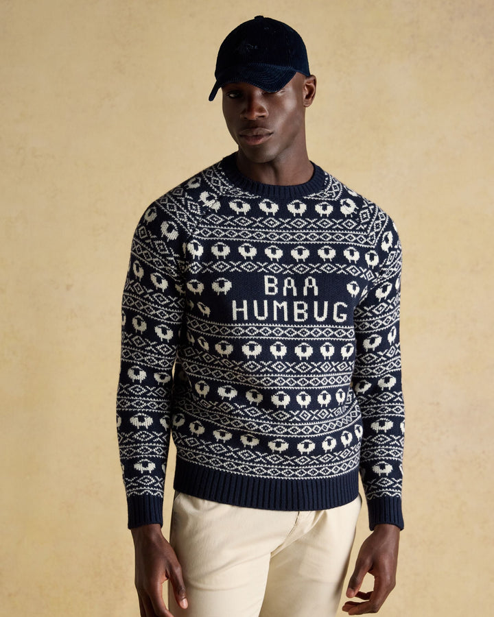 Man wearing a navy blue sweater with white sheep pattern and 'BAA HUMBUG' text, beige pants, and black cap on beige background