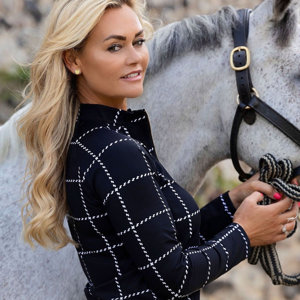 A blonde woman in a black equestrian long-sleeve shirt with a white stitched grid pattern stands beside a gray horse outdoors.