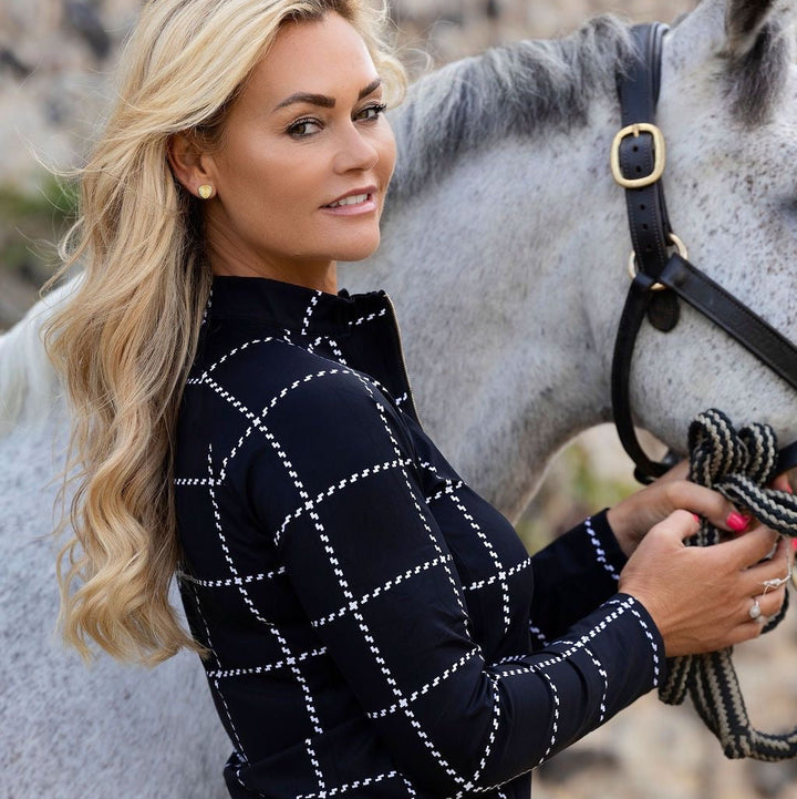 A blonde woman in a black equestrian long-sleeve shirt with a white stitched grid pattern stands beside a gray horse outdoors.