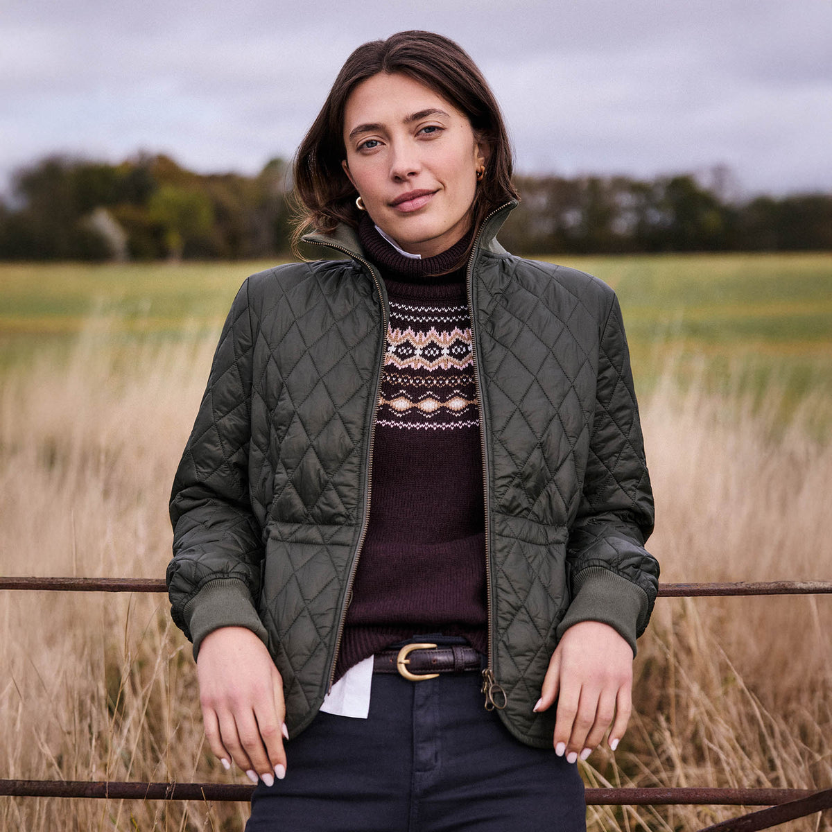 Woman leaning against a fence in a field wearing new Barbour clothing