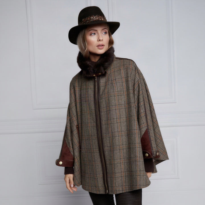 A woman wearing a brown and beige plaid poncho-style cape with dark brown faux-fur trim, a dark brown wide-brim hat, and black pants, standing against a light gray paneled wall.